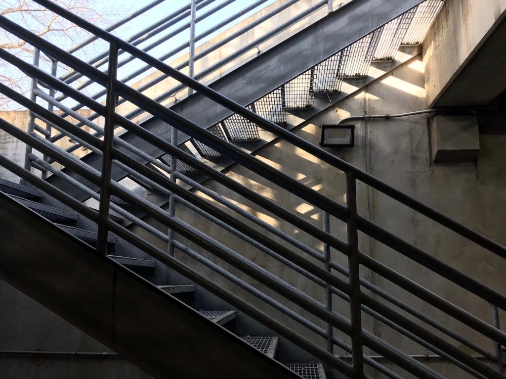 Crisscrossed open rail staircase, two flights, set into an alcove at the end of a parking garage, with a deep contrast between light and shadow
