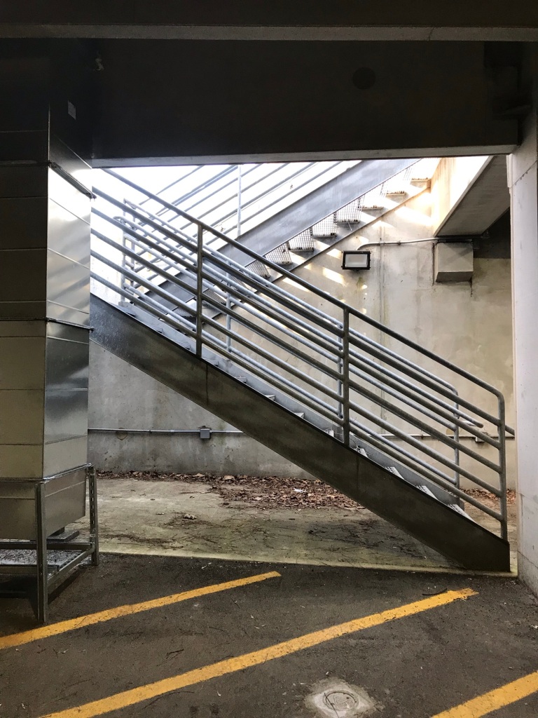 Crisscrossed open rail staircases in an alcove at the end of a parking garage, with diagonally painted yellow parking lines at the bottom